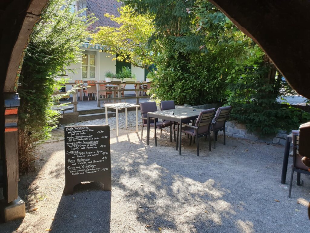 Außensitzbereich eines Cafés mit Tischen und Stühlen auf einem Kiesboden, einer Speisekarte auf einer schwarzen Tafel auf der linken Seite und Grünflächen rund um den Raum.