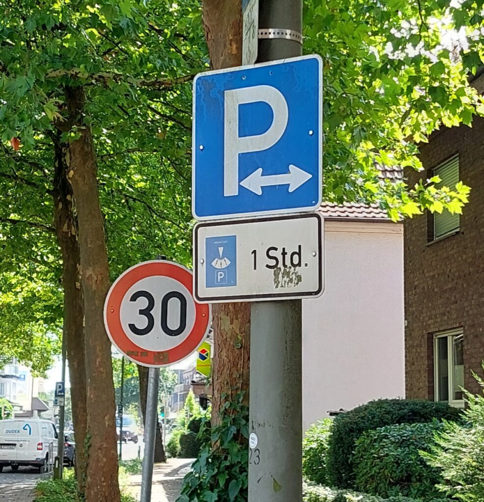 Ein blaues Parkschild mit nach links und rechts zeigenden Pfeilen zeigt eine Parkdauer von 1 Stunde (1 Std.) an. Im Hintergrund ist neben einem Baum und Gebäuden ein Schild mit einer Geschwindigkeitsbegrenzung von 30 km/h zu sehen.