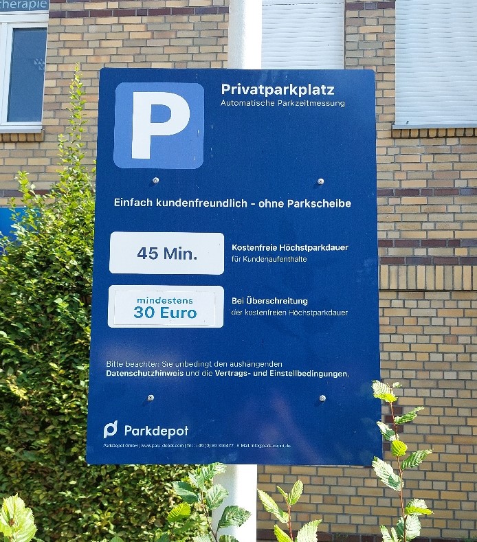 Schild mit Hinweis auf einen privaten Parkplatz mit automatischer Parkuhr. Es weist auf eine kundenfreundliche Gratiszeit von 45 Minuten hin, bei Überschreitung wird eine Mindestgebühr von 30 Euro erhoben. Befindet sich in der Nähe eines Backsteingebäudes.