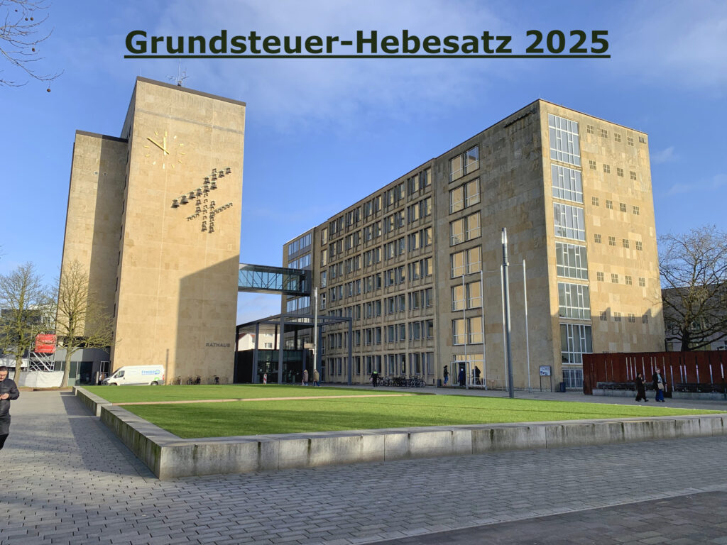 Ein großes Gebäude mit einem Uhrenturm und einem Verbindungsbau. Oben ist der Text „Grundsteuer-Hebesatz 2025“ zu sehen. Im Vordergrund sind eine Rasenfläche und ein Gehweg zu sehen.