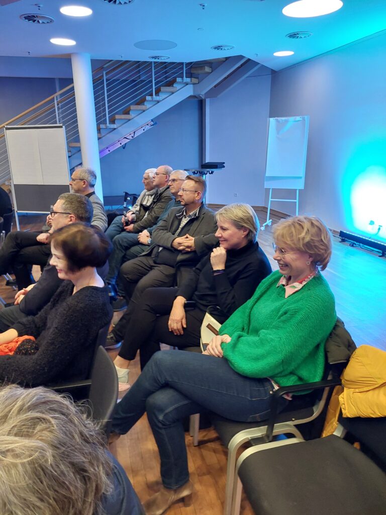Das Publikum sitzt im Innenbereich und nimmt an einer Veranstaltung mit in Reihen angeordneten Stühlen teil. Im Hintergrund sind eine Treppe und eine Beleuchtungsanlage zu sehen.