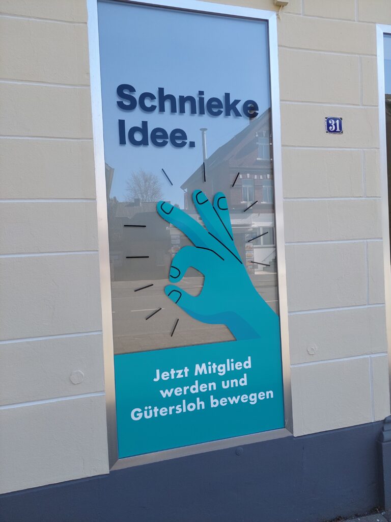 Auf einem Fensterschild ist eine blaugrüne Hand zu sehen, die eine „Okay“-Geste macht. Text lautet: „Schnieke Idee. Jetzt Mitglied werden und Gütersloh bewegen.“.