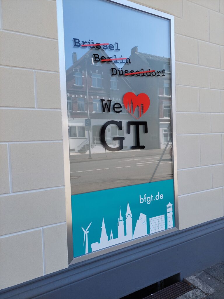 In einem Schaufenster sind die Namen von Brüssel, Berlin und Düsseldorf durchgestrichen, mit dem fettgedruckten Schriftzug „We ♥ GT“. Darunter sind Silhouetten von Sehenswürdigkeiten und die URL „bfgT.de“ zu sehen.