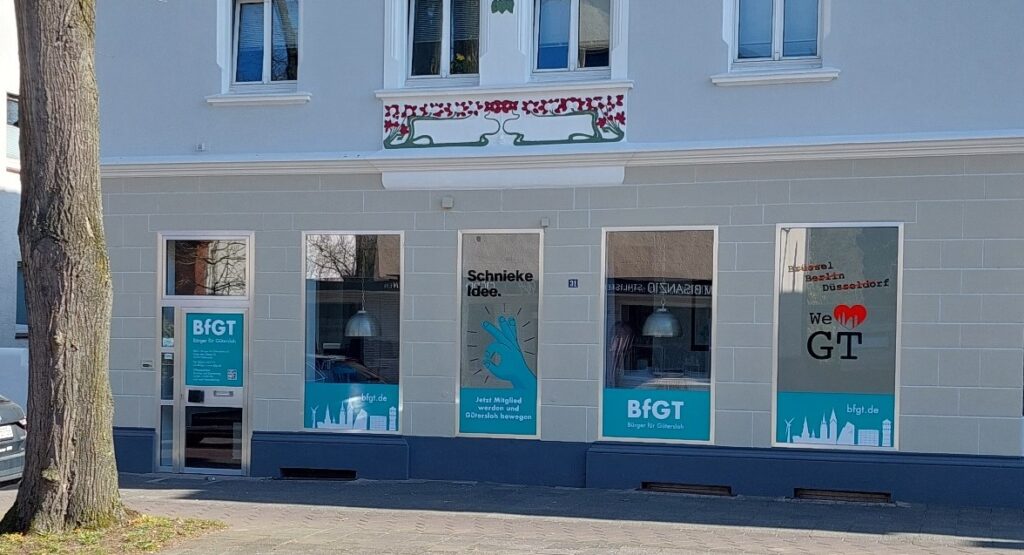 Schaufenster mit blauen „Bürger für Gütersloh“-Plakaten in den Fenstern, die ein Handsymbol und die Skyline der Stadt zeigen. Baum und Gehweg sind sichtbar. Das Gebäude hat oben eine dekorative Verzierung.