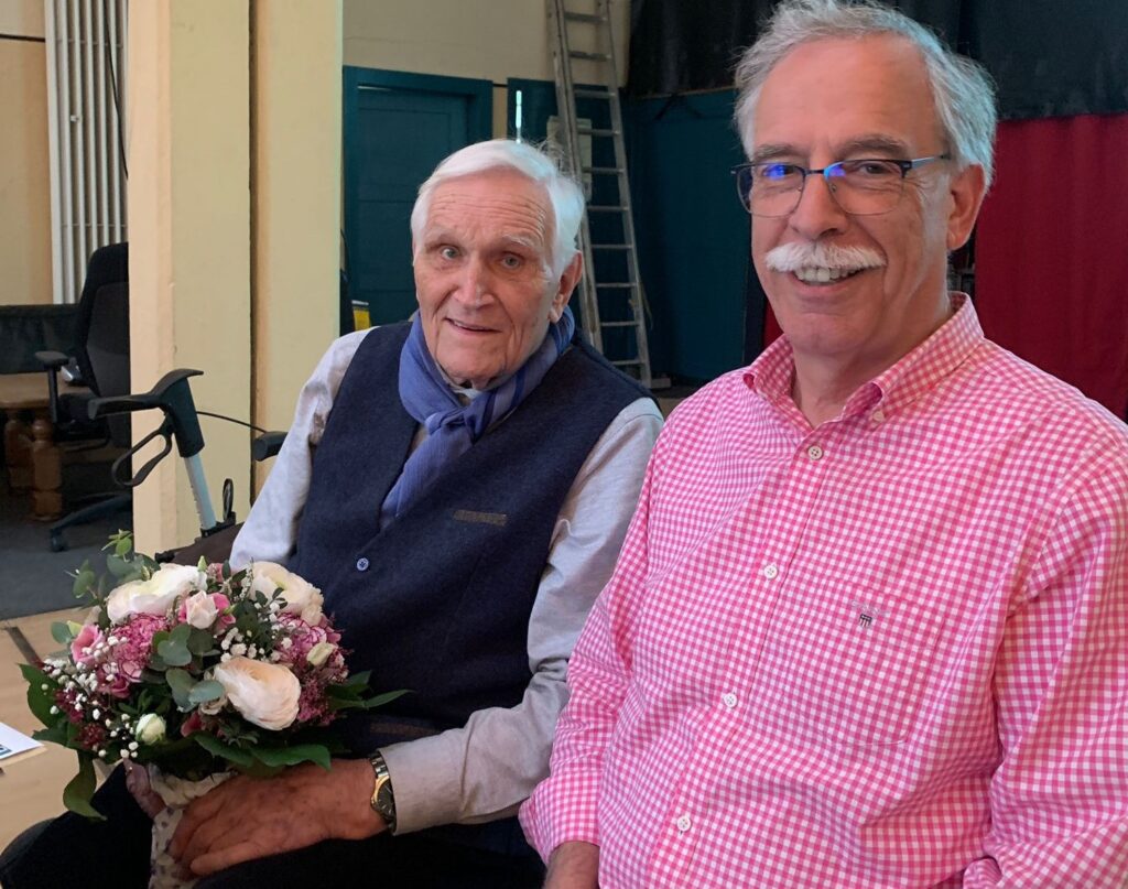 Zwei ältere Männer sitzen im Innenbereich nebeneinander; einer hält einen Blumenstrauß und trägt einen blauen Schal, der andere trägt eine Brille und ein rosa kariertes Hemd.