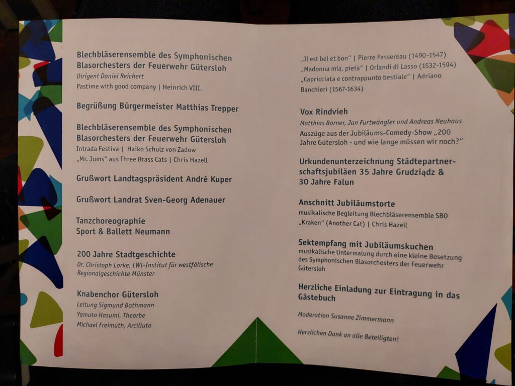 Programmbroschüre mit buntem abstrakten Rand, in der musikalische Darbietungen, Redner und Veranstaltungen für eine Feier aufgelistet sind, darunter Orchester, Chor, Tanzgruppe und offizielle Begrüßungsworte.