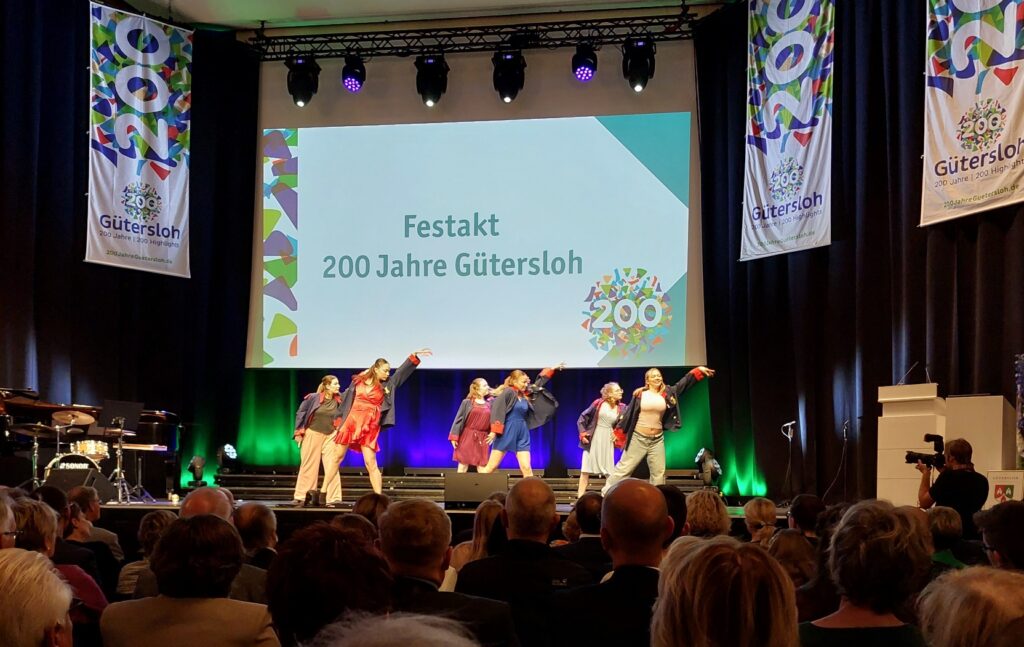 Bei einer Veranstaltung zum 200-jährigen Jubiläum der Stadt Gütersloh tritt eine Gruppe von Tänzern auf der Bühne auf. Das Publikum schaut zu und auf Transparenten ist das Jubiläumslogo zu sehen.
