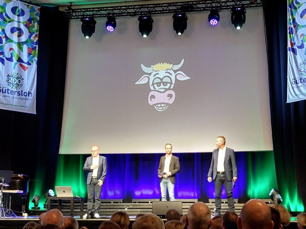Drei Männer stehen auf einer Bühne vor einer Leinwand, auf der ein Kuhgesicht im Cartoon-Stil zu sehen ist. Davor sitzt ein Publikum, und auf beiden Seiten sind Veranstaltungsbanner angebracht.