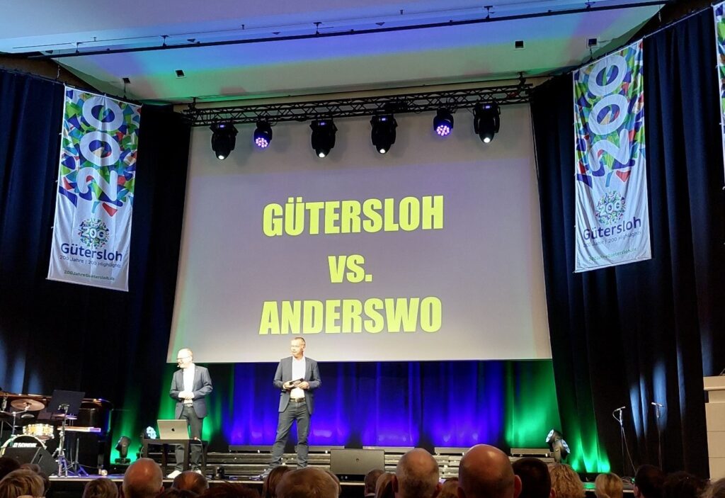 Bei einer Veranstaltung stehen zwei Männer auf einer Bühne vor einer Leinwand mit der Aufschrift „Gütersloh vs. Anderswo“, im Hintergrund sind Transparente und bunte Beleuchtung zu sehen.