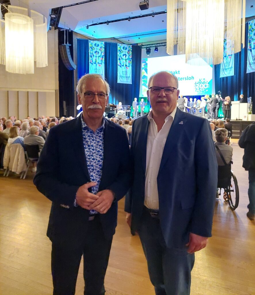 Zwei Männer in Anzügen stehen in einem großen Auditorium mit sitzendem Publikum und einer Bühne im Hintergrund, auf der sich Menschen unter hellem Licht versammelt haben.