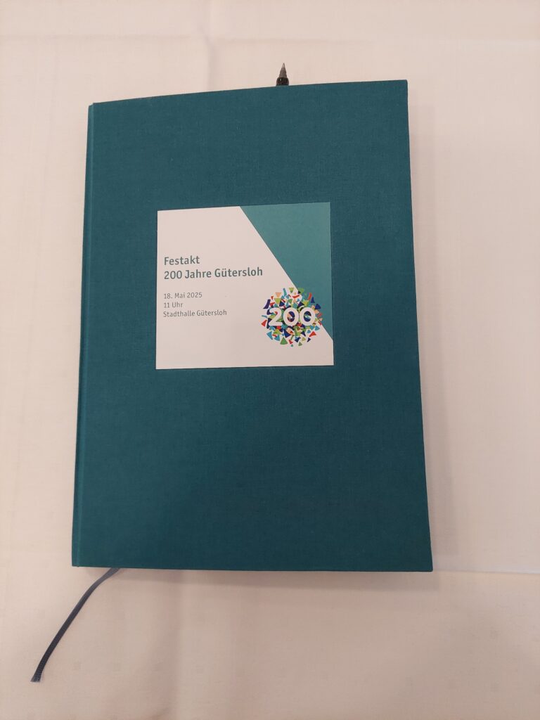 Ein blaugrünes Buch mit der Aufschrift „Festakt 200 Jahre Gütersloh, 18. Mai 2025, 11 Uhr, Stadthalle Gütersloh“ und einer farbenfrohen „200“-Grafik, das auf einer weißen Oberfläche liegt.