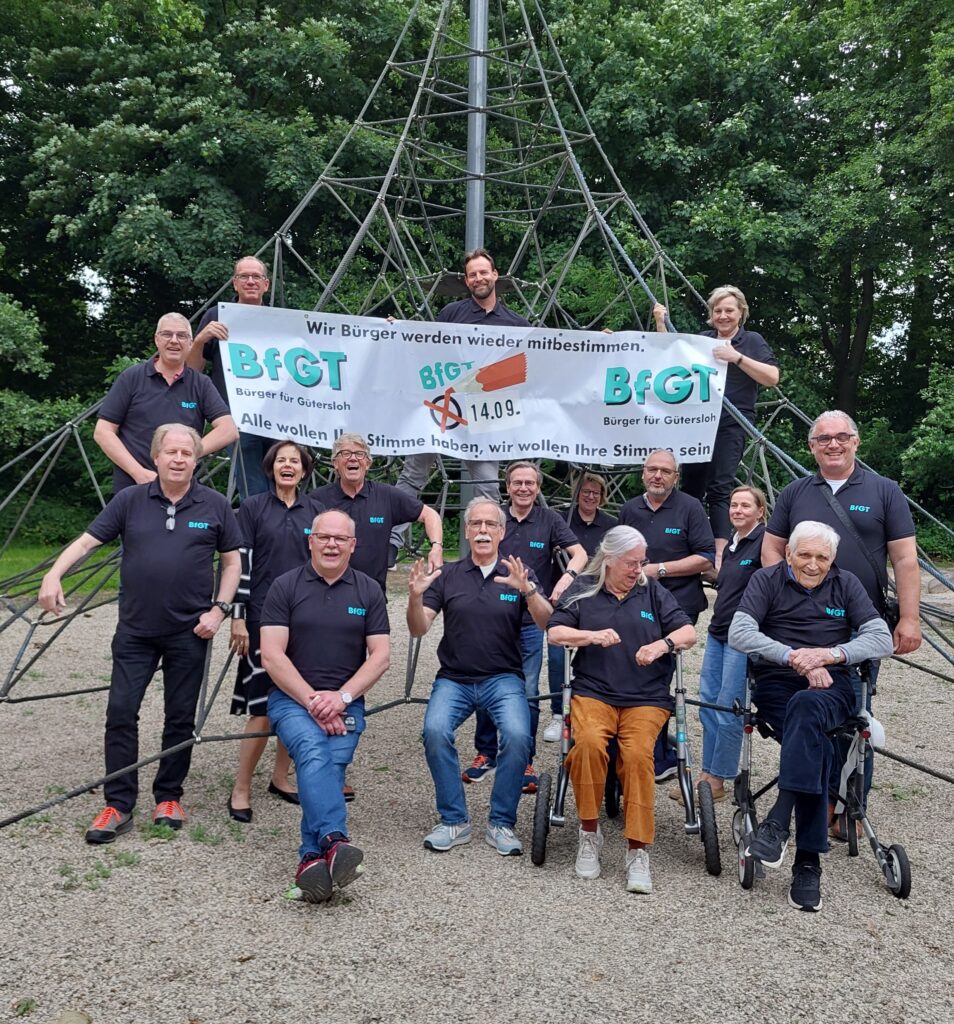 Eine Gruppe von Menschen in schwarzen Hemden posiert vor einer Kletterseilstruktur und hält ein Banner mit der Aufschrift „BfGT“ und einem deutschen Text hoch. Im Freien sind Bäume im Hintergrund zu sehen.