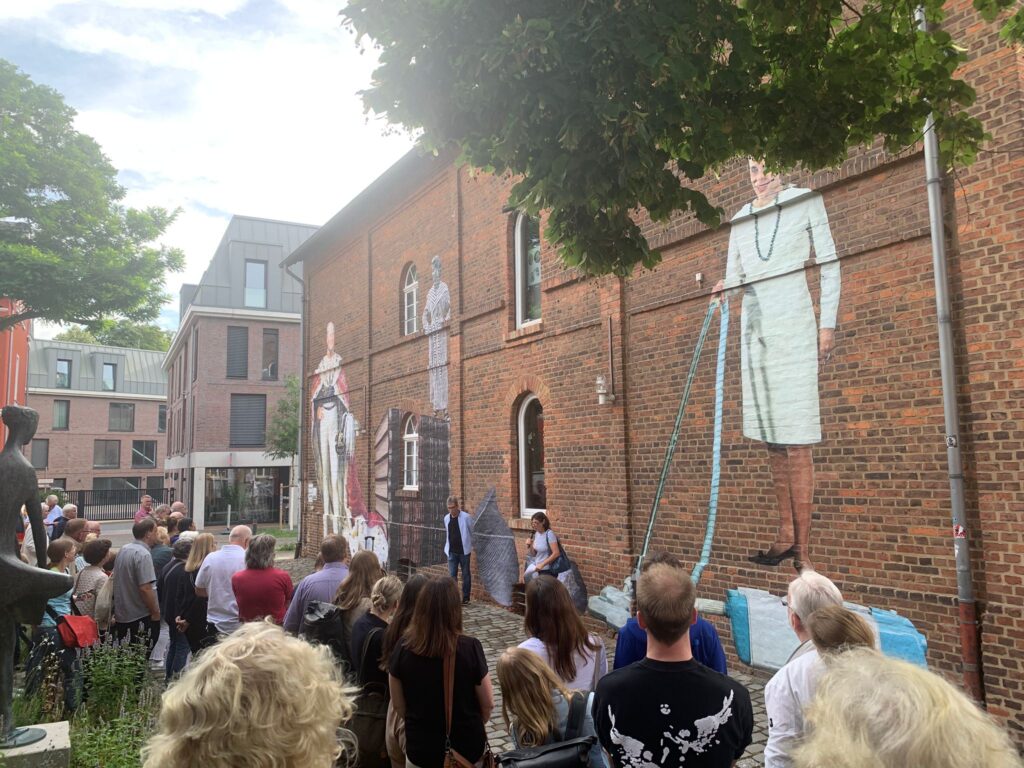Eine Menschenmenge versammelt sich in einem Innenhof und lauscht den Lautsprechern vor einem Backsteingebäude mit großen Wandmalereien von Menschen an der Wand.