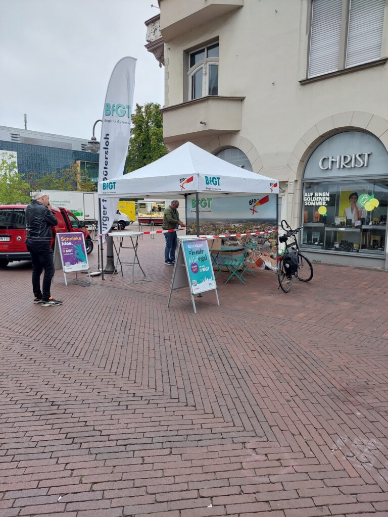 Ein weißes Werbezelt mit Informationsschildern steht auf einem gemauerten Gehweg in der Nähe eines Geschäfts. Zwei Personen sind anwesend, und ein Fahrrad ist in der Nähe geparkt.