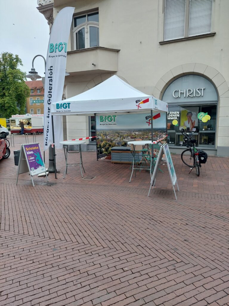 Ein Werbestand mit einem weißen Zelt, Bannern und Schildern ist auf einem Backsteinweg vor einem Geschäft mit dem Namen "Christ" aufgebaut. In der Nähe ist ein Fahrrad geparkt.