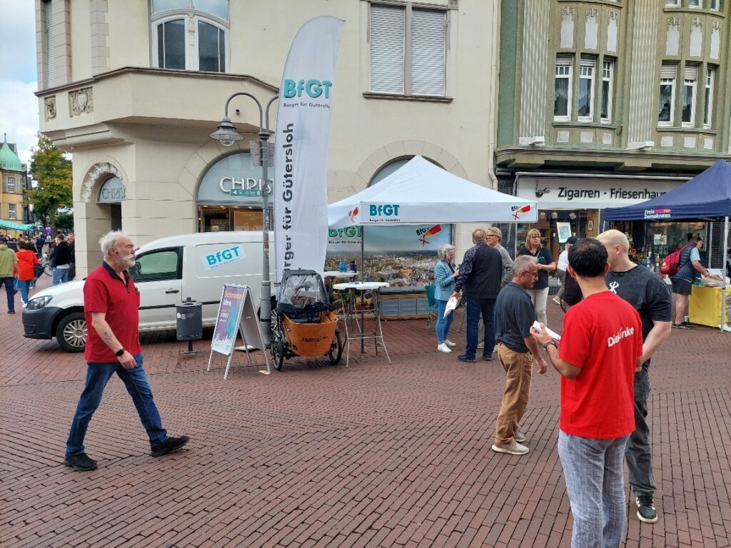 Straßenszene mit Menschen in der Nähe von Informationsständen und Bannern, geparkten Lieferwagen und historischen Gebäuden im Hintergrund.