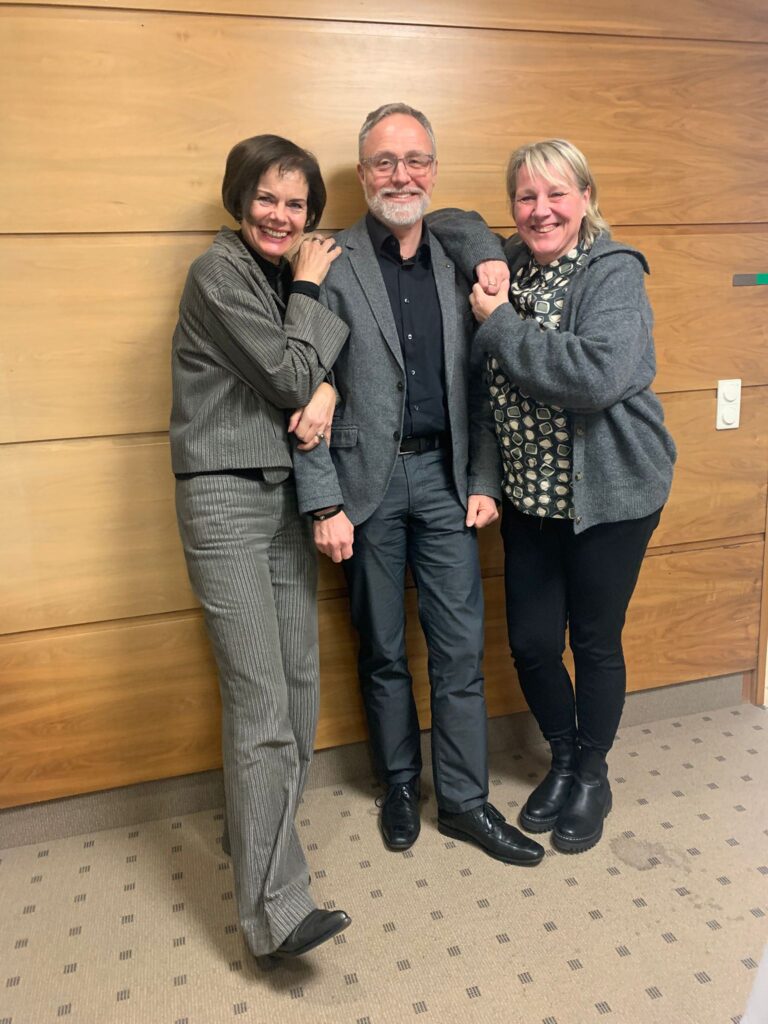 Drei Erwachsene stehen lächelnd drinnen an einer Holzwand; zwei Frauen stehen auf beiden Seiten eines Mannes, alle in Business-Casual-Kleidung gekleidet.