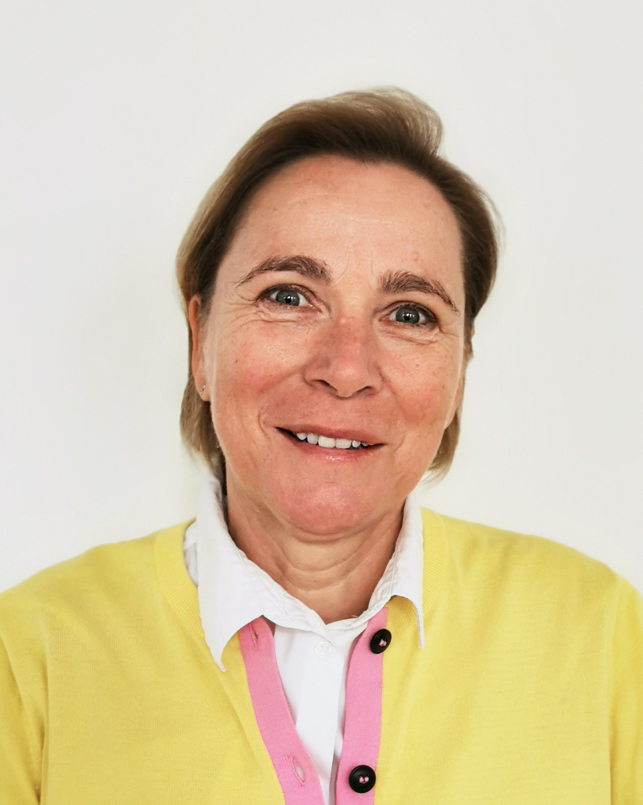 Eine Frau mittleren Alters mit kurzen hellbraunen Haaren lächelt in die Kamera. Sie trägt einen gelben Pullover über einem weißen Hemd mit Kragen und einem rosa Akzent. Der Hintergrund ist einfarbig weiß.