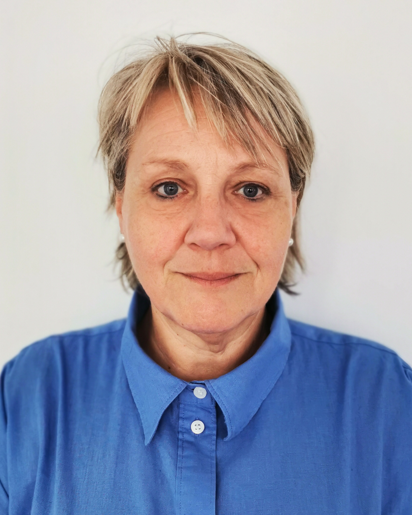 Eine Person mittleren Alters mit kurzen blonden Haaren und blauem Kragenhemd steht vor einem schlichten weißen Hintergrund und schaut mit neutralem Blick direkt in die Kamera.