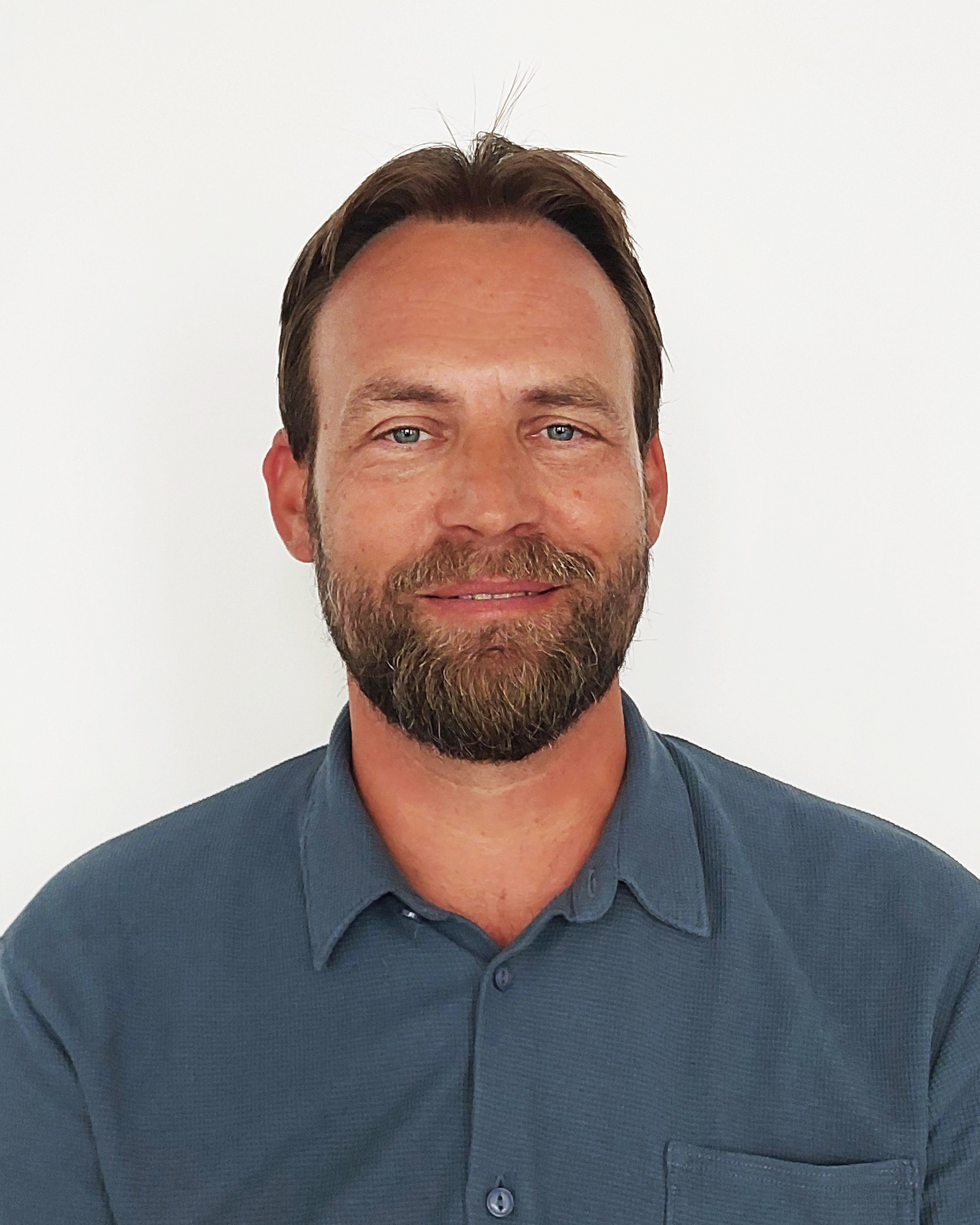 Ein Mann mit braunem Haar und Bart, der ein blaues Hemd mit Kragen trägt, lächelt in die Kamera vor einem schlichten weißen Hintergrund.