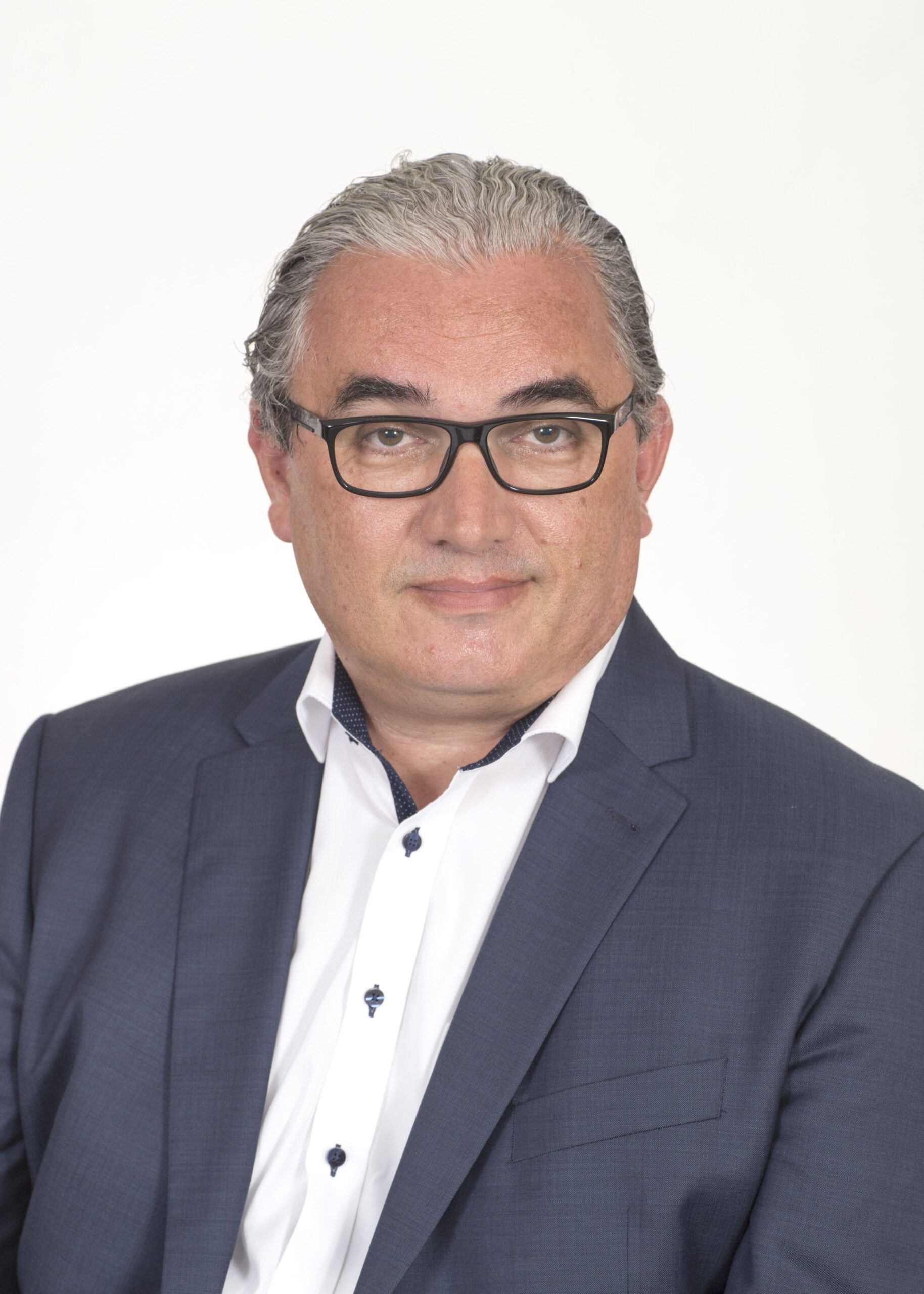Ein Mann mittleren Alters mit grauem Haar und Brille, der einen marineblauen Anzug und ein weißes Hemd trägt, posiert vor einem schlichten weißen Hintergrund.