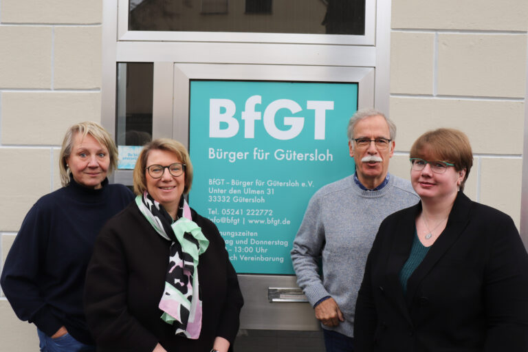 Vier Erwachsene stehen vor einer Tür mit einem Schild mit der Aufschrift "Bürger für Gütersloh" und den Kontaktdaten der Organisation BfGT.