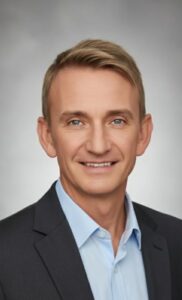 Ein Mann mittleren Alters mit kurzen blonden Haaren, der eine dunkle Anzugsjacke und ein hellblaues Hemd trägt, lächelt in die Kamera vor einem schlichten grauen Hintergrund.