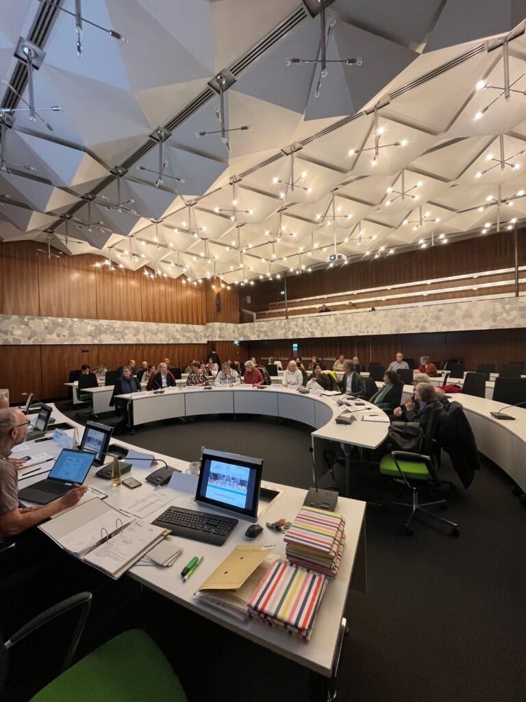 Menschen sitzen um einen großen geschwungenen Konferenztisch in einem modernen Konferenzraum mit Computern, Notebooks und Deckenbeleuchtung.