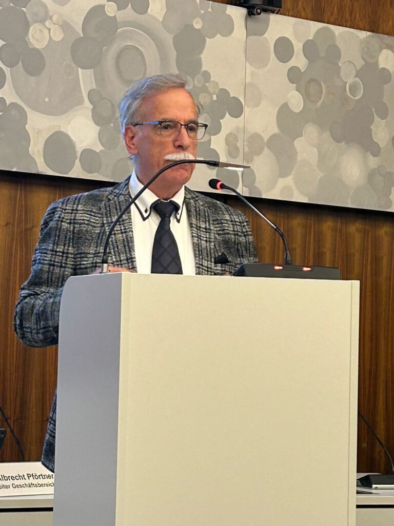 Ein Mann in Anzug und Krawatte steht an einem Podium mit einem Mikrofon und spricht in einer formellen Umgebung mit gemusterten Wandpaneelen hinter ihm.