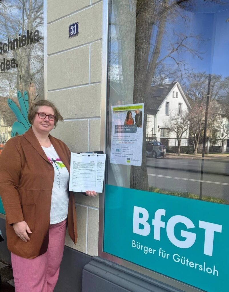 Eine Person steht vor einem Gebäude mit Dokumenten in der Hand, neben einem Fenster mit der Aufschrift "BfGT Bürger für Gütersloh".