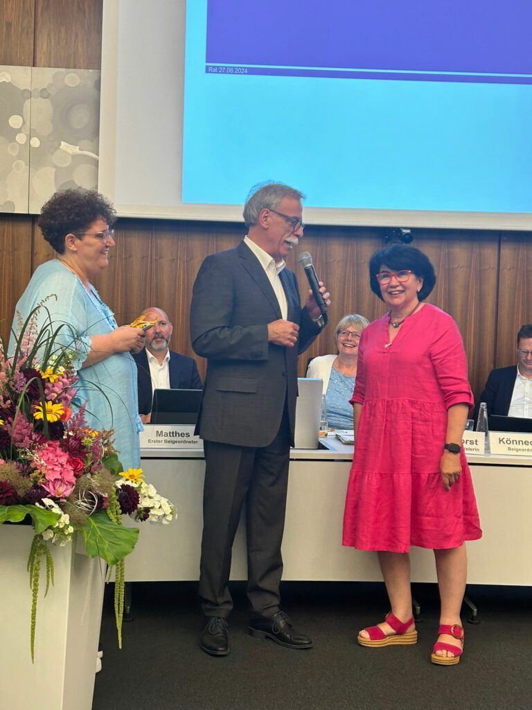 Eine Frau in einem rosa Kleid steht da und lächelt, während ein Mann im Anzug in ein Mikrofon spricht. Eine weitere Frau in einem blauen Outfit steht in der Nähe und hält einen Notizblock. Auf einem Tisch links sind Blumen arrangiert.