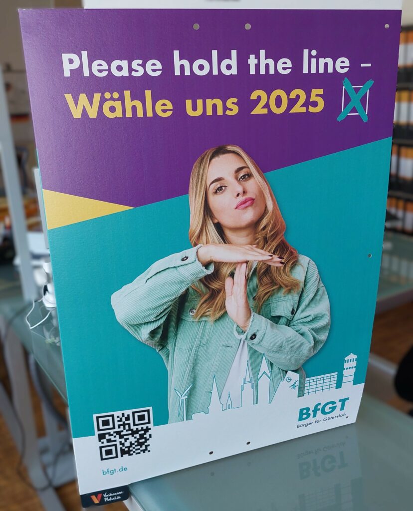 Ein Plakat mit einer Frau, die eine „Auszeit“-Geste macht, dem Text „Bitte bleiben Sie am Apparat – Wähle uns 2025“ und „BFGT“ mit einem QR-Code.