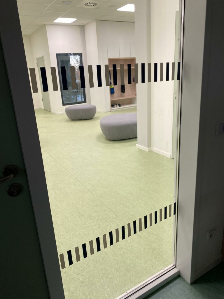 Ein Raum mit hellgrünem Boden und zwei grauen ovalen Bänken. Die Tür hat eine Reihe schwarzer vertikaler Linien als Designelemente. Im Hintergrund sind Regale und ein Fenster zu sehen.
