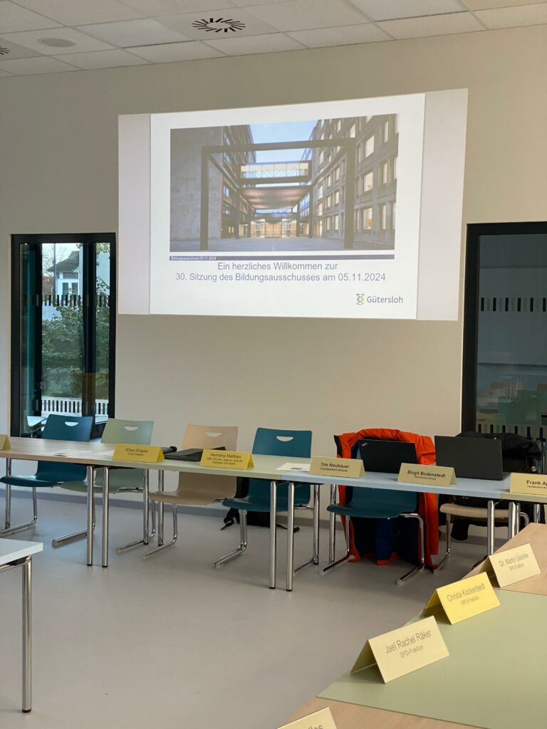 Konferenzraum mit Präsentationsbildschirm, auf dem Text und ein Architekturbild angezeigt werden. Stühle und Namensschilder sind um den Tisch herum angeordnet.