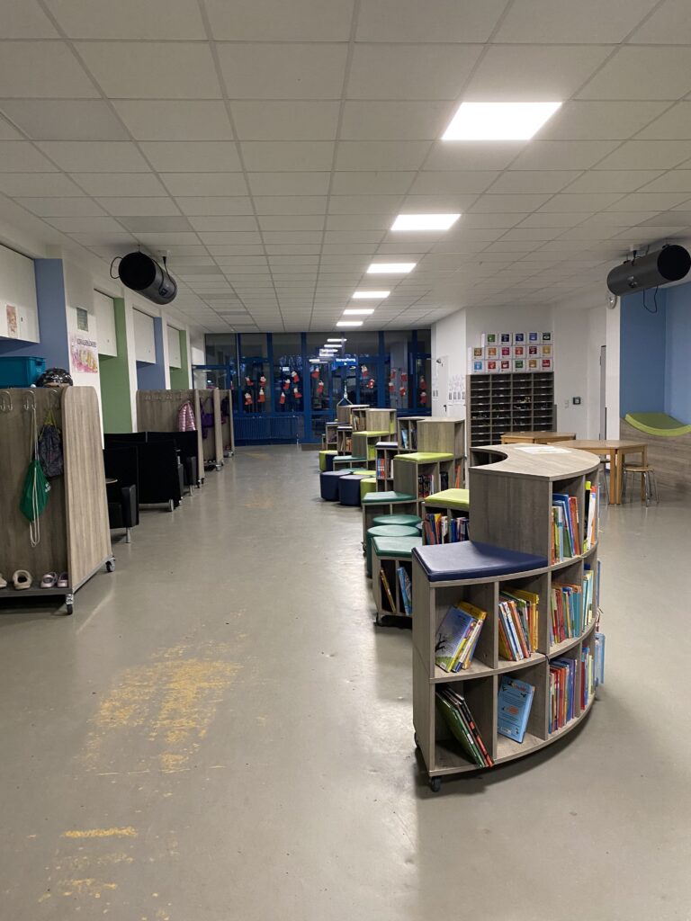 Ein Schulflur mit Bücherregalen und Hockern an den Wänden. Auf der linken Seite sind Haken mit Mänteln zu sehen, und am anderen Ende stehen Tische und Stühle. Leuchtstofflampen beleuchten den Bereich.
