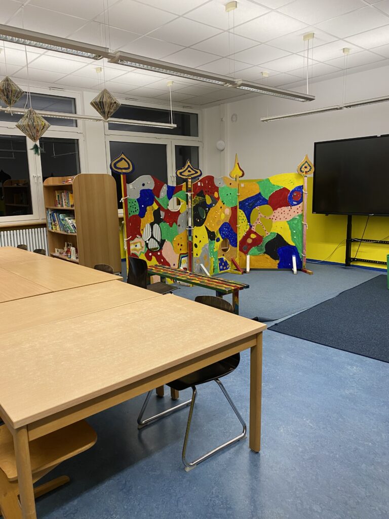 Klassenzimmer mit Tischen, Stühlen, Bücherregalen und einer bunten Trennwand. Großer Bildschirm im Hintergrund. Helle Deckenbeleuchtung.