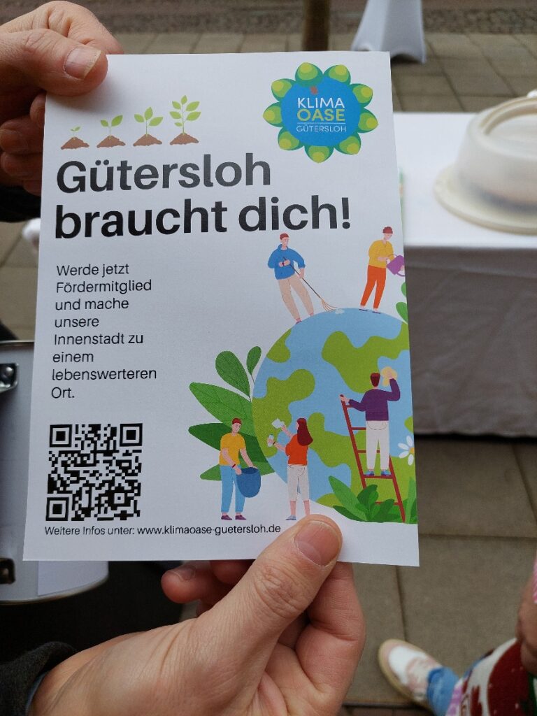 Person hält einen Flyer mit Abbildungen von Garten- und Malern. Der Text lautet: „Gütersloh braucht dich!“ mit einem QR-Code und einer Webadresse für weitere Informationen.
