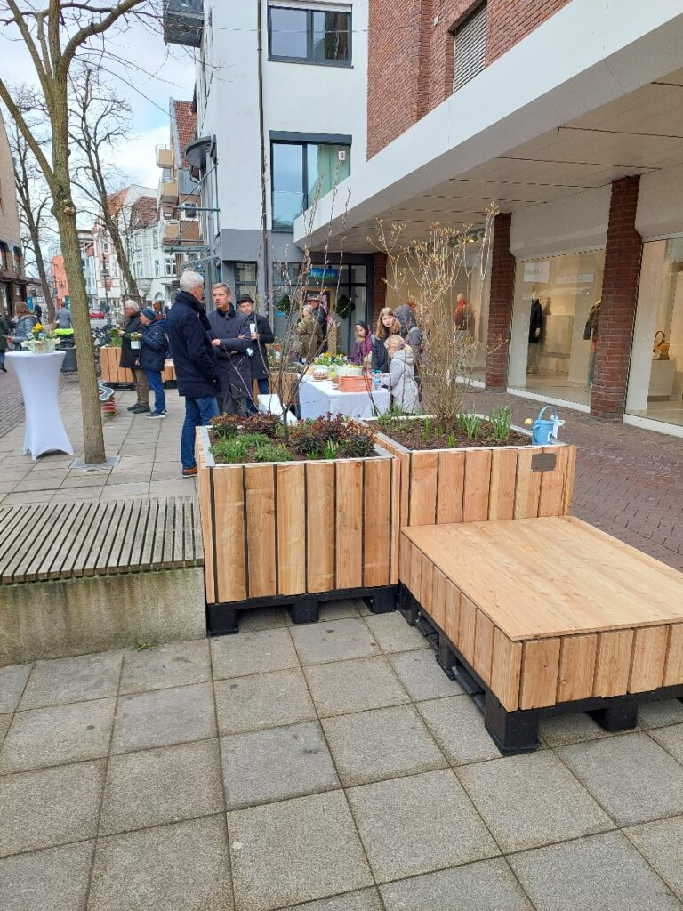 Menschen versammelten sich um einen Straßenmarktstand mit Pflanzen und Kunsthandwerk. In der Nähe stehen Holzbänke und Pflanzkübel. Im Hintergrund sind Gebäude zu sehen.