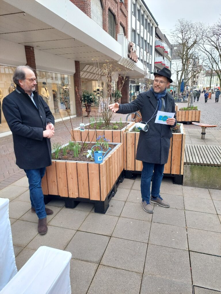 Zwei Männer stehen auf einem Bürgersteig neben Hochbeeten. Einer gestikuliert und hält Papiere in der Hand; der andere beobachtet. Im Hintergrund sind ein Gebäude und eine leere Straße zu sehen.