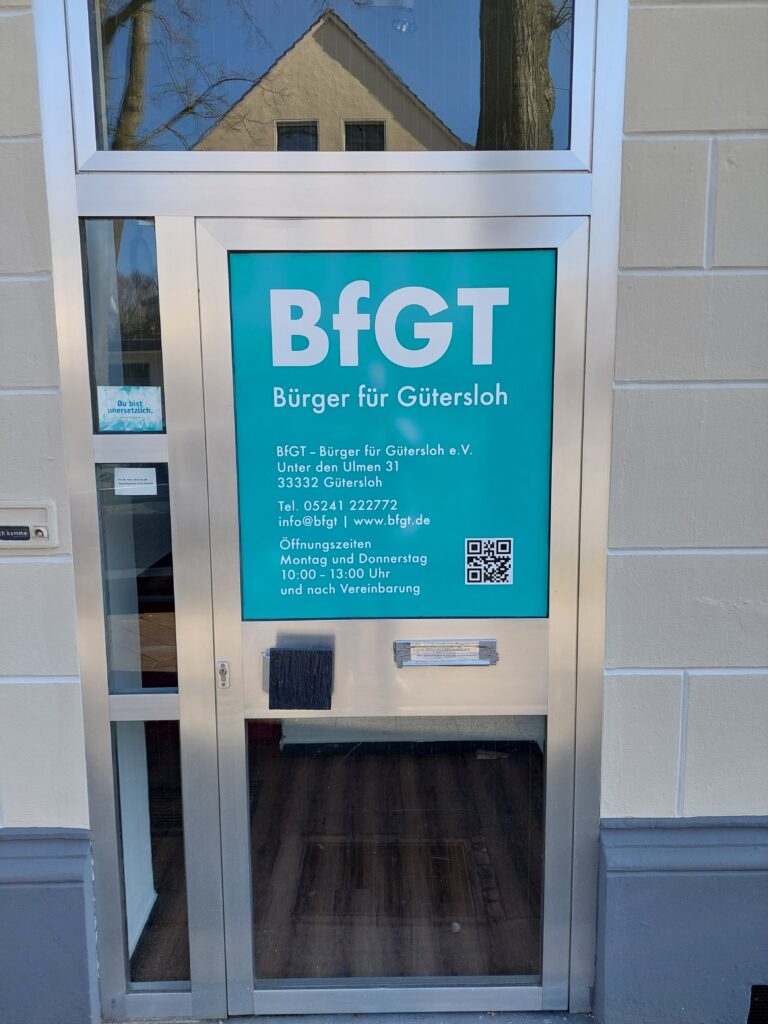 Eine Glastür mit einem blaugrünen Schild für „BfGT Bürger für Gütersloh“ mit Kontaktdaten, Öffnungszeiten und einem QR-Code. Auf der Tür spiegeln sich Bäume und ein Gebäude.