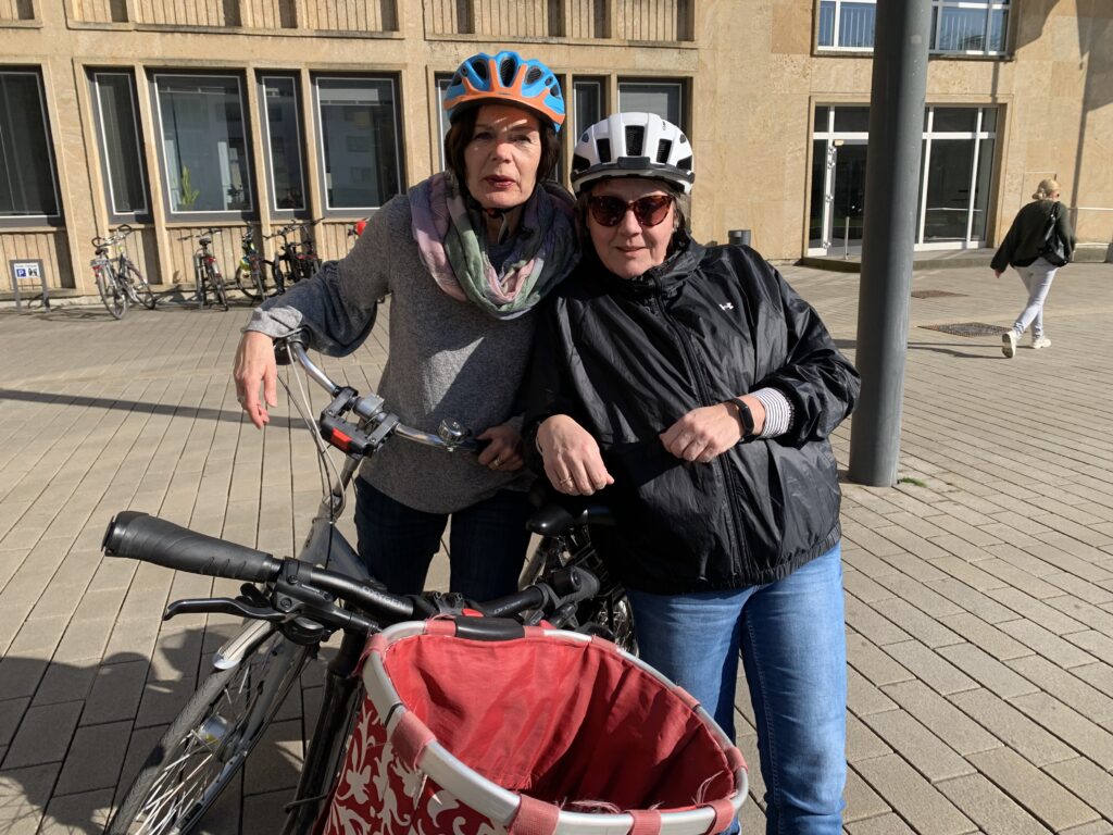 Zwei Personen mit Helmen stehen neben einem Fahrrad mit einem roten Korb auf einer gepflasterten Fläche mit Gebäuden und einigen geparkten Fahrrädern im Hintergrund.