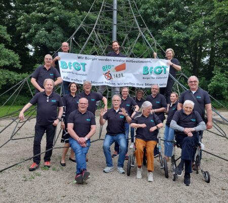 Eine Gruppe von Menschen, einige davon im Rollstuhl, posiert in einem Park vor einem Klettergerüst aus Metall und einem Banner der politischen Gruppe BFGT.