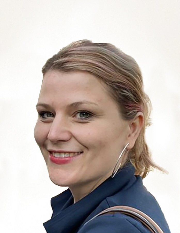 Eine Frau mit hellbraunem, nach hinten gestecktem Haar, die große Ohrringe und eine marineblaue Jacke trägt, lächelt in die Kamera vor einem schlichten, hellen Hintergrund.