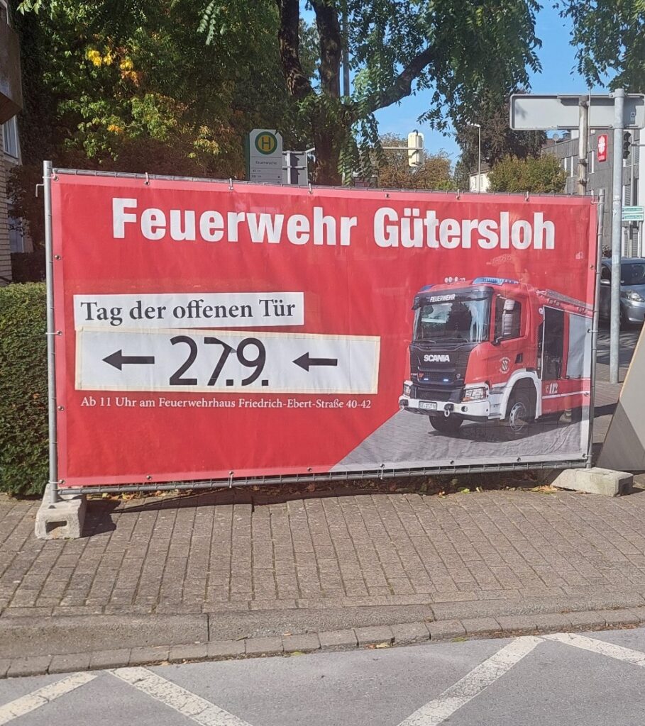 Rotes Werbebanner für den Tag der offenen Tür der „Feuerwehr Gütersloh“ am 27.9. mit dem Bild eines Feuerwehrautos, Veranstaltungsdetails und Angaben zum Veranstaltungsort auf einem Bürgersteig in der Stadt.