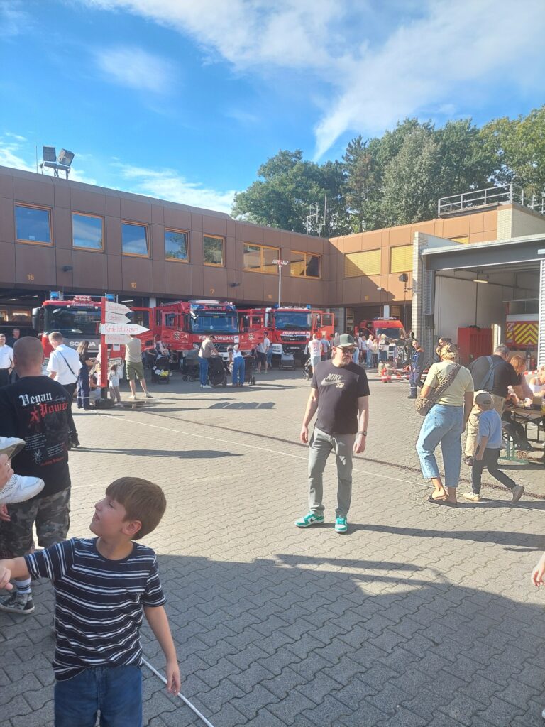 Vor einer Feuerwache, vor der mehrere Löschfahrzeuge geparkt sind, haben sich Menschen versammelt. Einige Erwachsene und Kinder gehen umher, während andere rechts an Tischen sitzen. Der Himmel ist teilweise bewölkt.