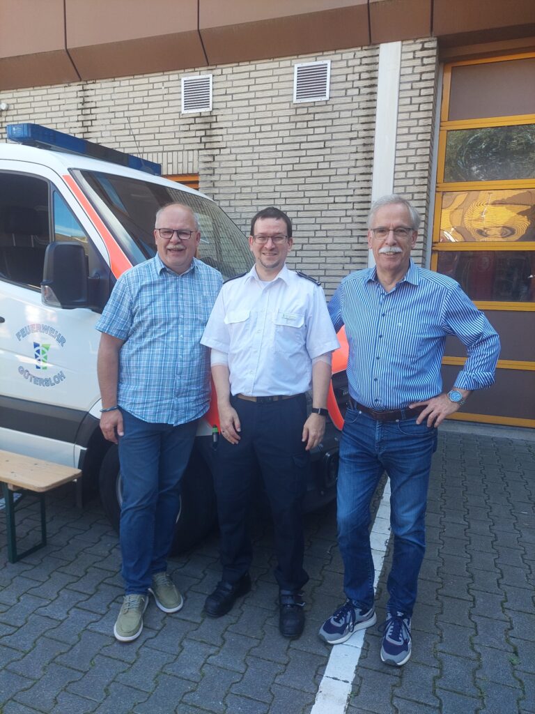 Drei Männer stehen lächelnd vor einem Einsatzfahrzeug, das neben einem Backsteingebäude geparkt ist. Der Mann in der Mitte trägt ein weißes Uniformhemd; die beiden anderen tragen karierte Hemden und Jeans.