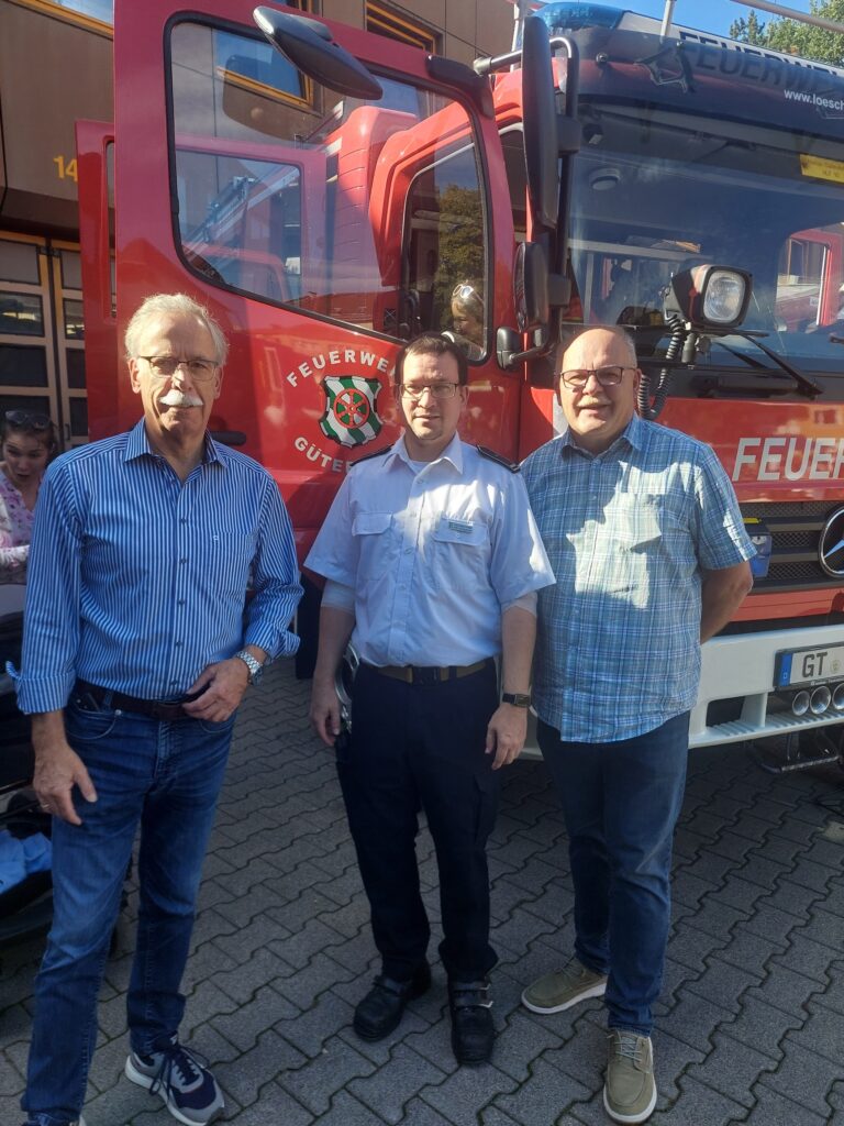 Drei Männer stehen vor einem roten Feuerwehrauto; der Mann in der Mitte trägt eine Feuerwehruniform, während die beiden anderen Freizeitkleidung tragen.