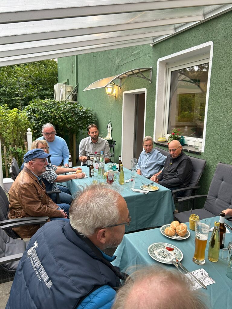 Acht Männer sitzen mit Getränken und Essen im Freien unter einer überdachten Terrasse an zwei Tischen und unterhalten sich. Die Atmosphäre wirkt ungezwungen und entspannt.