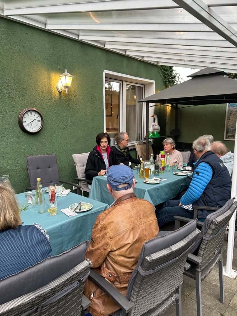 Acht Erwachsene sitzen im Freien unter einem Vordach um einen Tisch herum; auf dem Tisch stehen Getränke und Teller, daneben befindet sich eine grüne Wand mit einer Uhr.